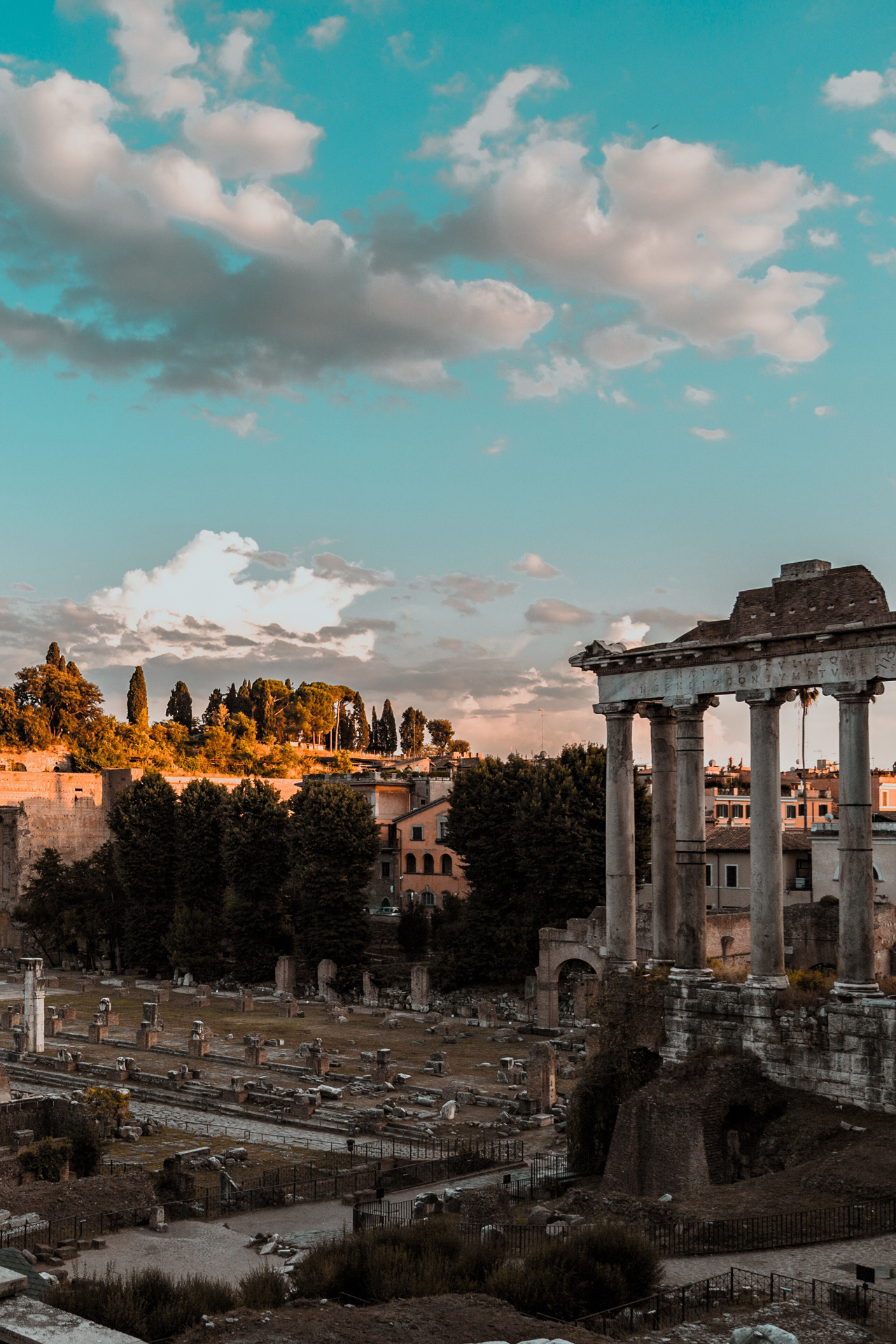 Ancient Roman ruins (the Fori Romani) with stone columns and scattered remains, with trees and buildings in the background