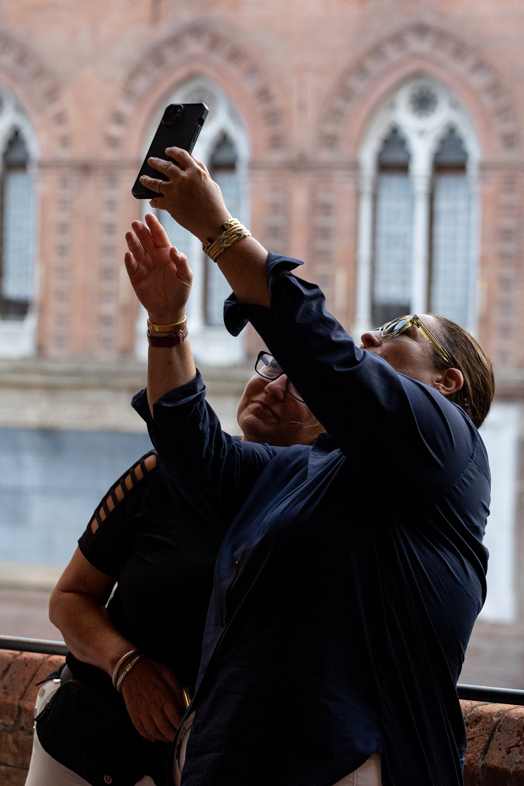 Two people taking a selfie with a smartphone in front of a brick building with arched windows.