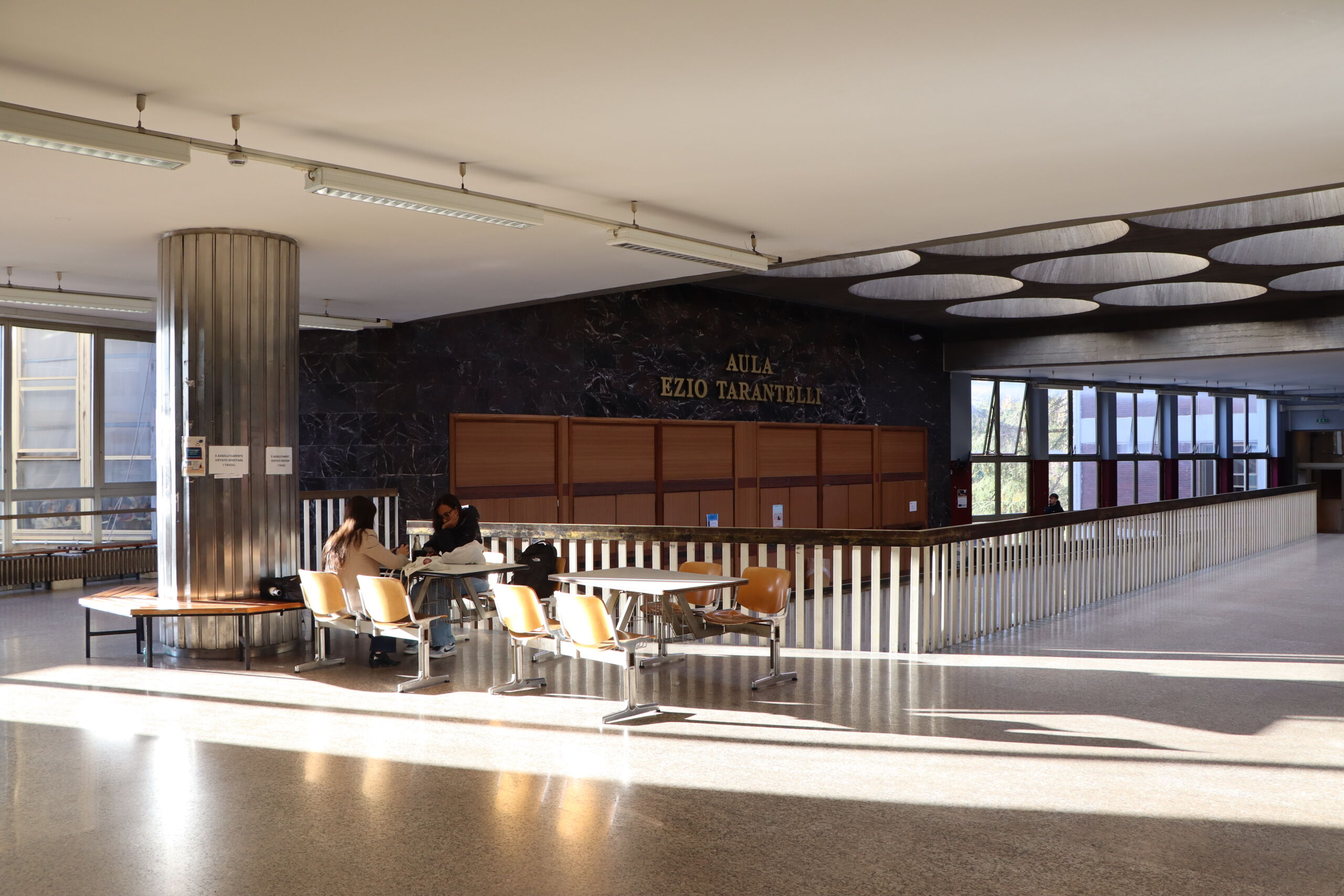 Interior view of a university hallway with seating area and students studying near the entrance to Aula Ezio Tarantelli