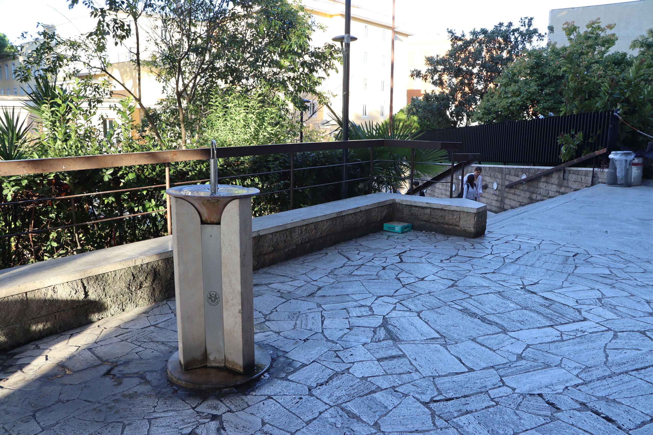 Outdoor area with a drinking fountain, stone pavement, and stairs surrounded by greenery