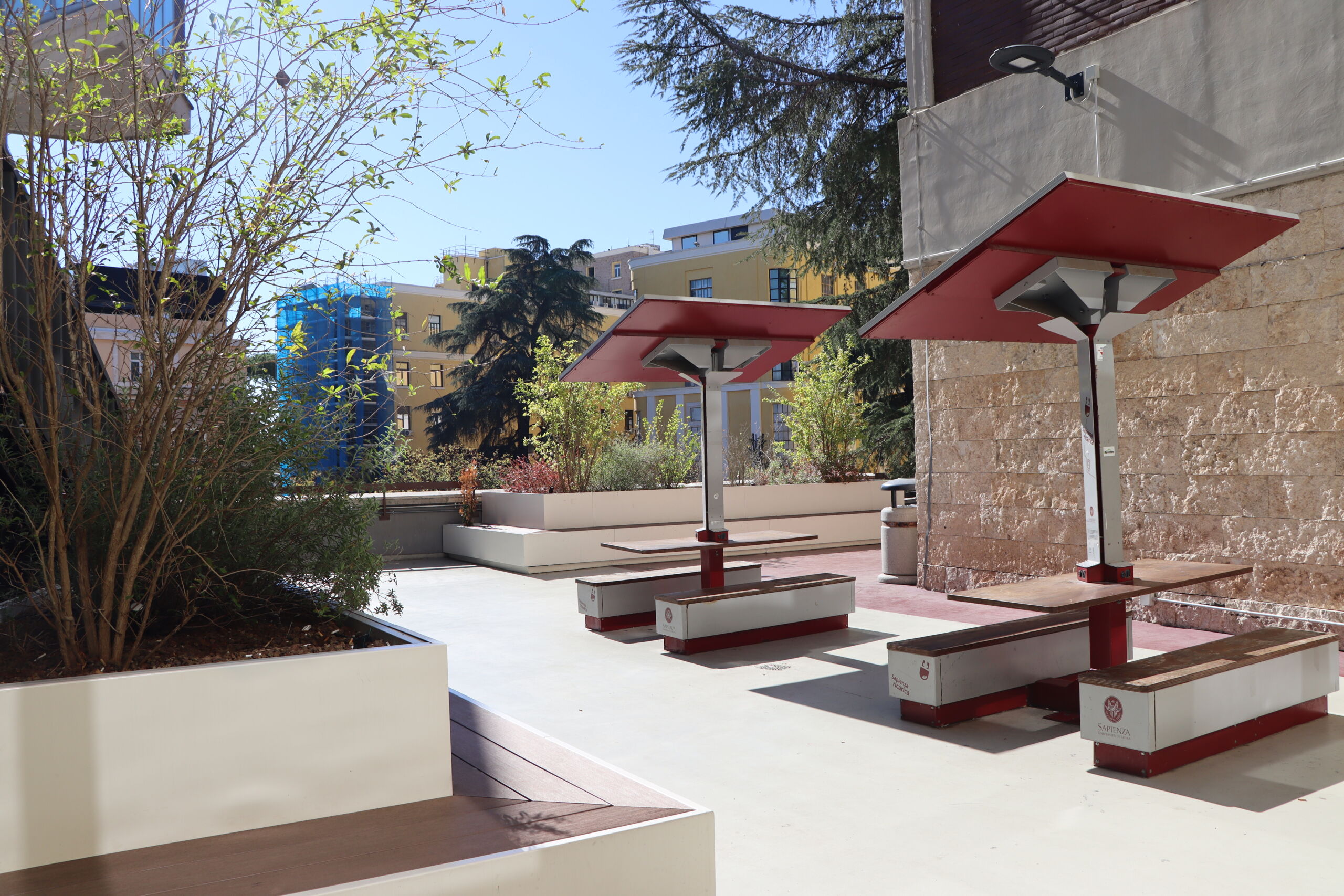 Outdoor seating area with modern benches and tables under red canopies, surrounded by planters and nearby buildings