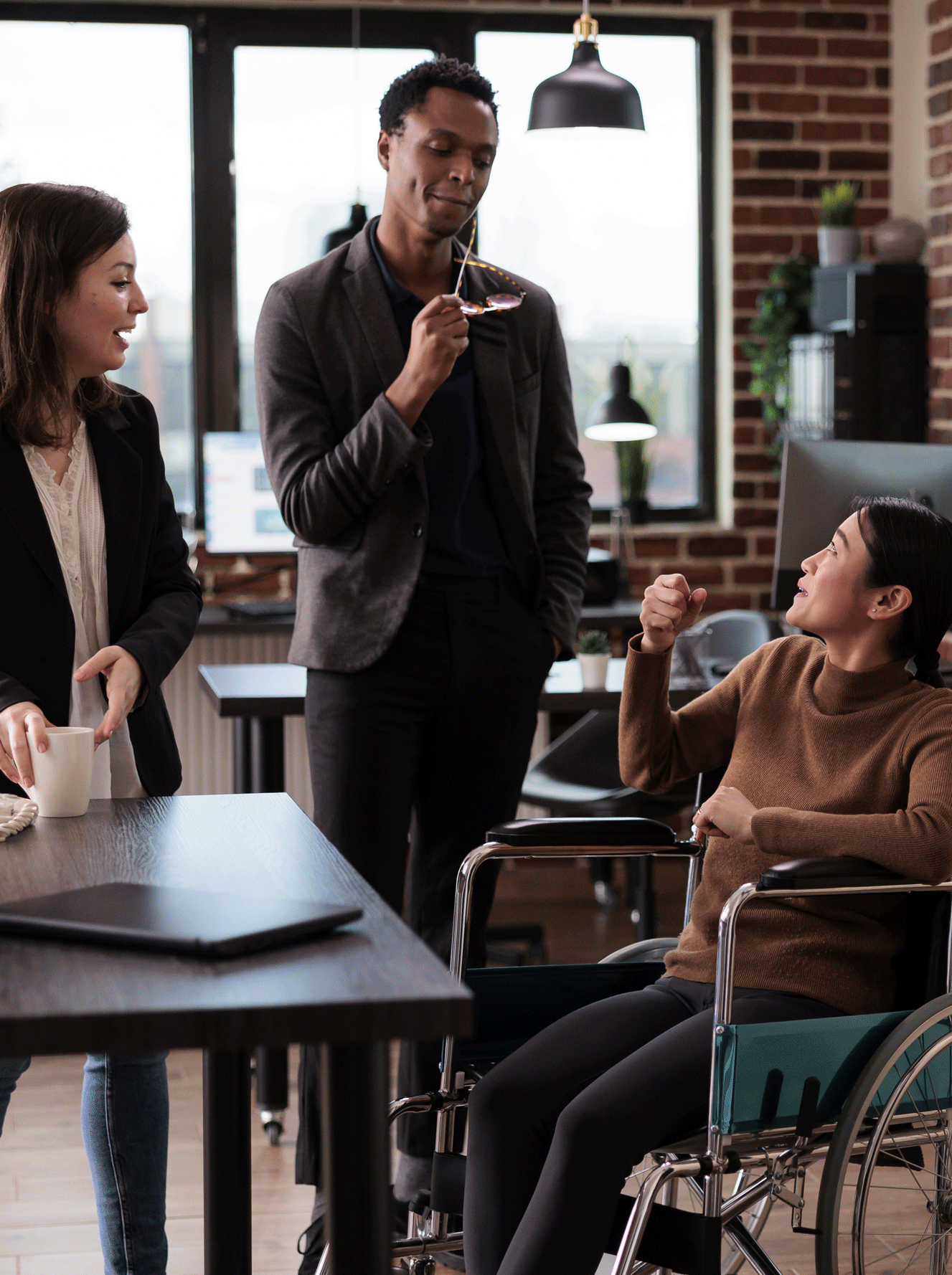 Three colleagues having a discussion in a modern office. One white woman is holding a coffee mug, a black man in a suit is standing with glasses in hand, and an asian woman in a wheelchair is engaging in conversation with them. They appear to be collaborating in a casual work setting with laptops and office furniture visible.